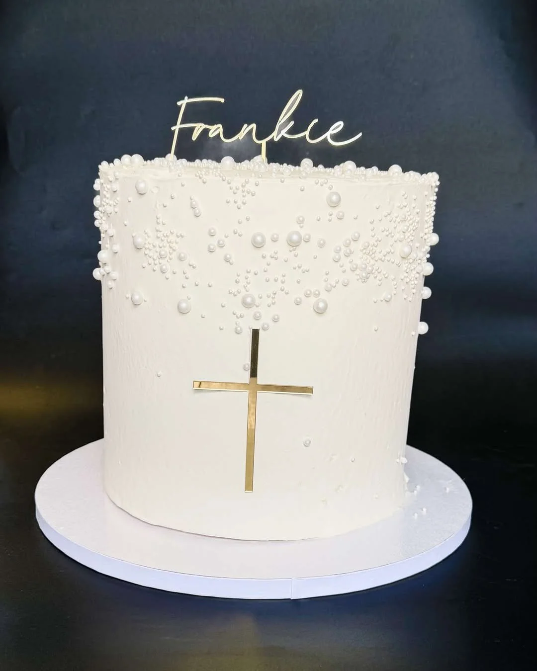 A white cylindrical cake decorated with pearl-like edible beads, a gold cross on the front, and a gold script name "Frankie" on top.