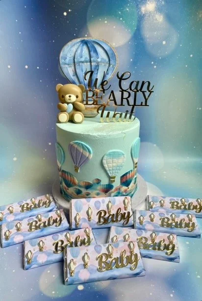 A baby shower cake with hot air balloon decorations, teddy bear topper, and a sign that says 'We Can Bearly Wait.' Surrounded by boxes labeled 'Baby' with hot air balloon designs.