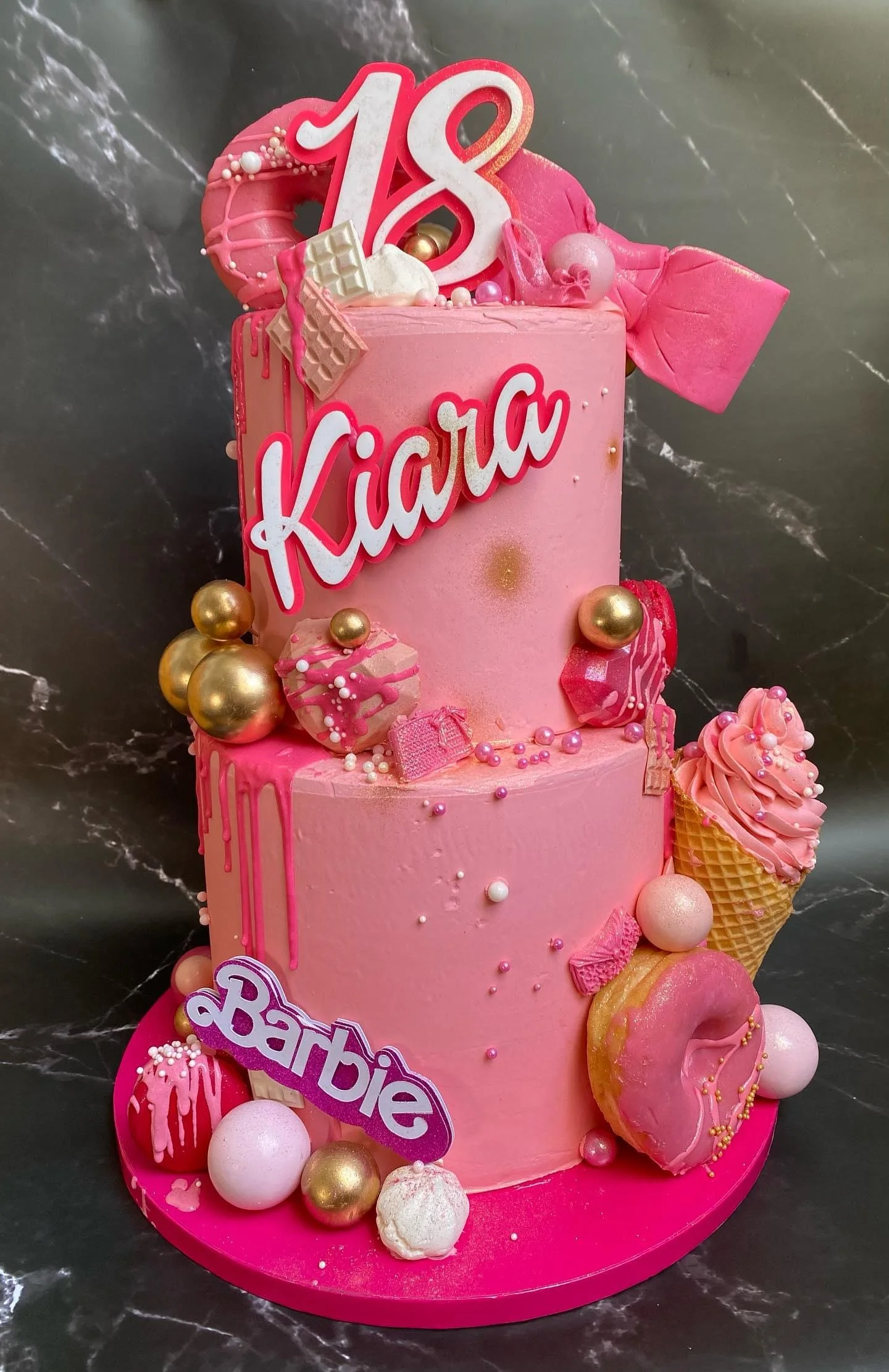 Pink birthday cake decorated with a large white and red '18' topper, the name 'Kiarra' in pink letters, and various pink, white, and gold decorations including candies and balls, sitting on a pink cake board.