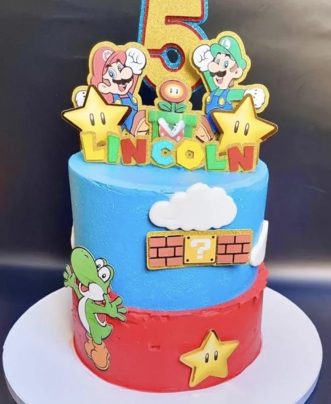 Colorful birthday cake with Super Mario theme decorations. It has two tiers, the top tier is blue with a white cloud and brick block with a yellow question mark, and the bottom tier is red with a yellow star. There are cartoon images of Mario, Luigi,