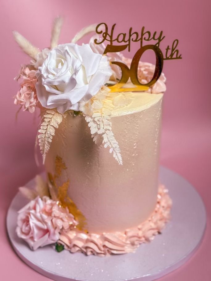 A pink 50th birthday cake decorated with white and pink flowers and a gold "Happy 50th" cake topper, set against a pink background.