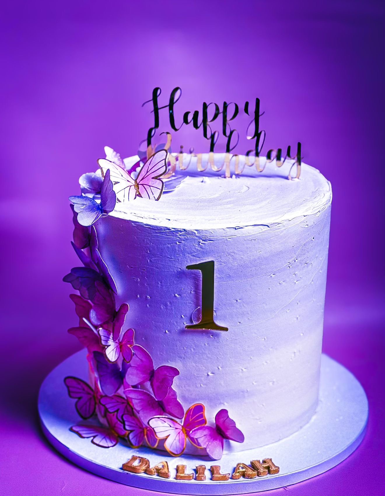 White birthday cake decorated with purple butterfly toppers, purple orchids, a black 'Happy Birthday' topper, a gold number '1', and the name 'DALLAH' in gold letters on the cake board, set against a purple background.