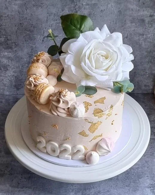 A white cake decorated with a large white rose and green leaves, adorned with gold leaf accents, macarons, meringues, and other small confections, with "2022" written on the side.