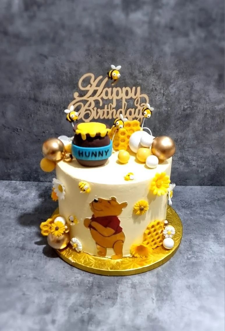 A birthday cake decorated with honey bees, flowers, and a Winnie the Pooh illustration, featuring a "Happy Birthday" topper with bee accents.
