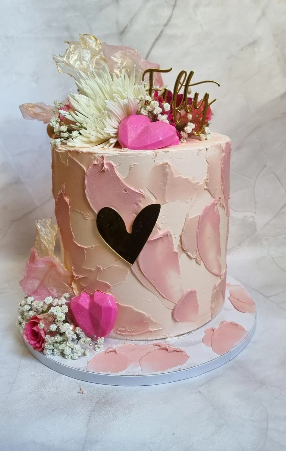 A pink frosted birthday cake decorated with flowers, a black heart, pink hearts, and a gold "Happy" topper. The cake is on a white base with pink petal-like decorations around it.