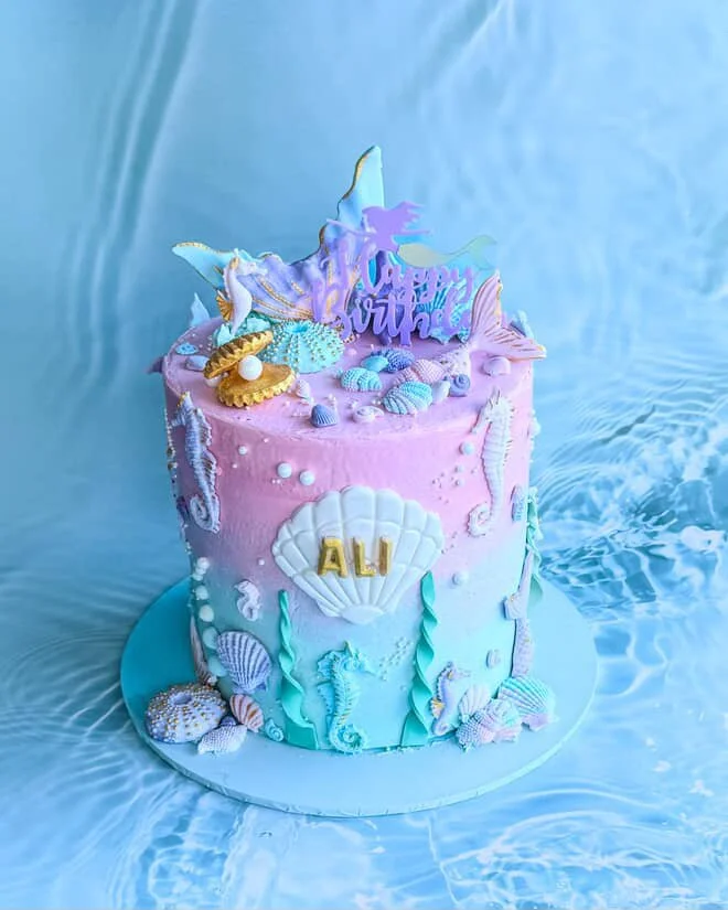 Sea-themed birthday cake with pink and blue icing, seashell decorations, starfish, coral, and a "Happy Birthday" topper, personalized for Ali.