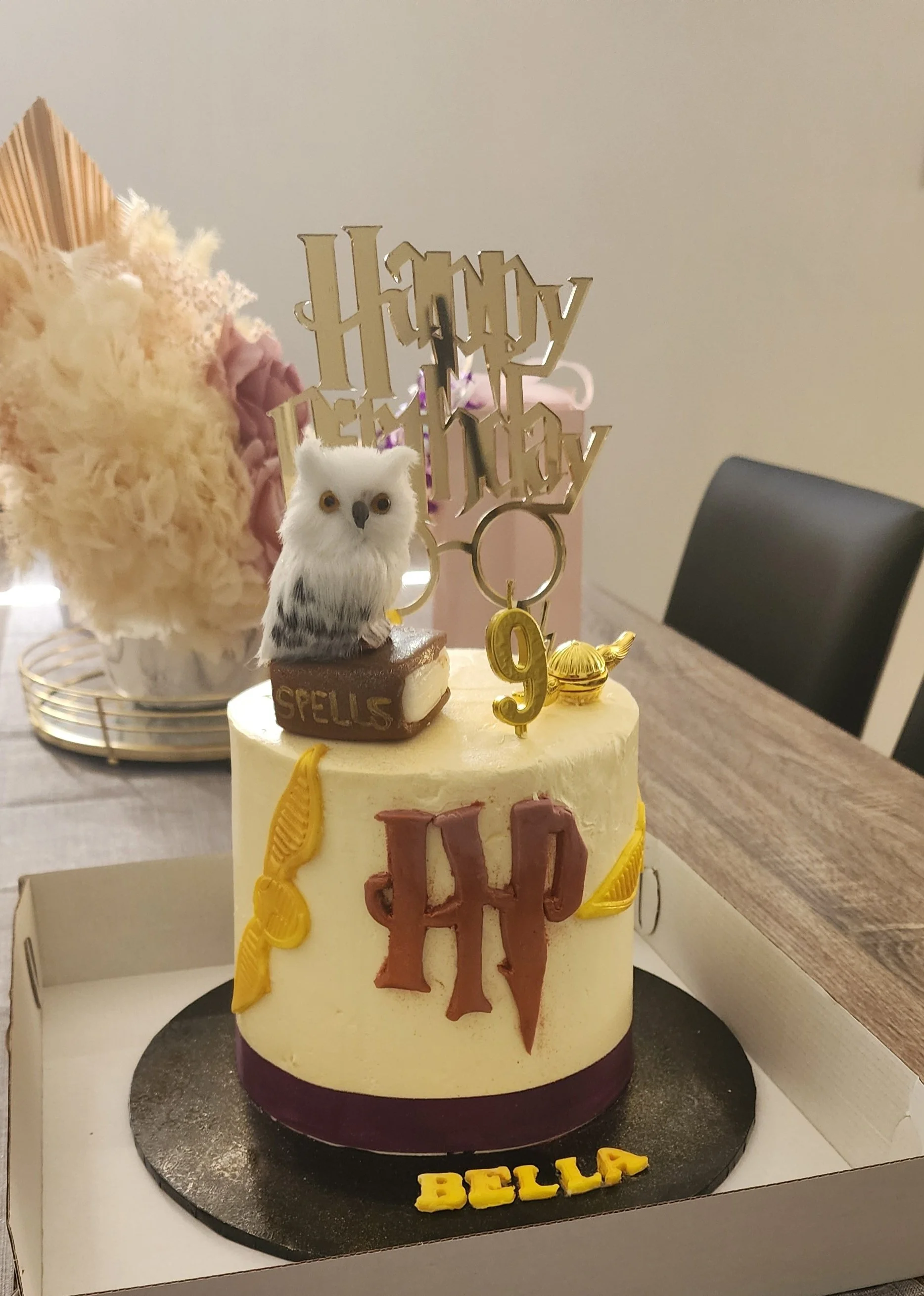 A birthday cake with woodland animal decorations, including a plush owl, and gold and chocolate letters spelling 'BELLA' with additional 'Happy Birthday' topper and yellow ribbon decorations. The cake is in a white box on a table, with a blurred back