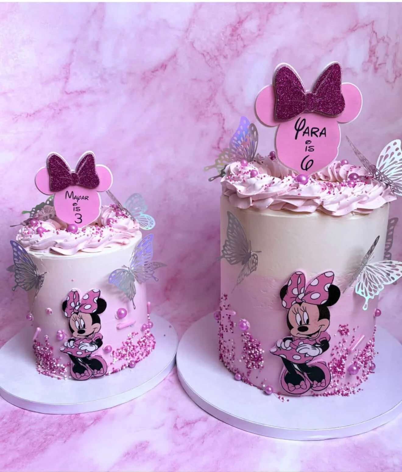 Two pink Minnie Mouse-themed birthday cakes decorated with pink sprinkles, purple pearls, and butterfly-shaped edible decorations. Each cake has a Minnie Mouse figure with a pink polka-dot bow, and pink Minnie Mouse-shaped toppers with handwritten me