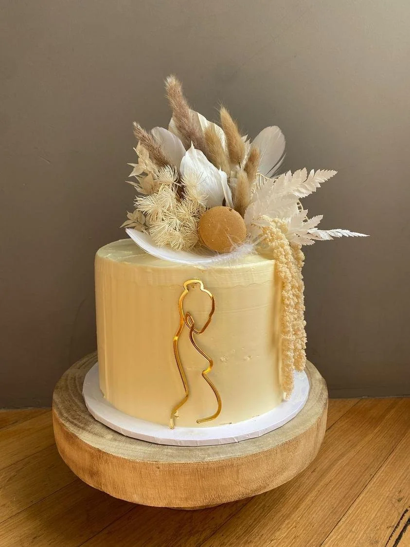 A cream-colored cake decorated with dried flowers, a gold cake topper of a woman's silhouette, and dried pampas grass, sitting on a wooden cake stand.