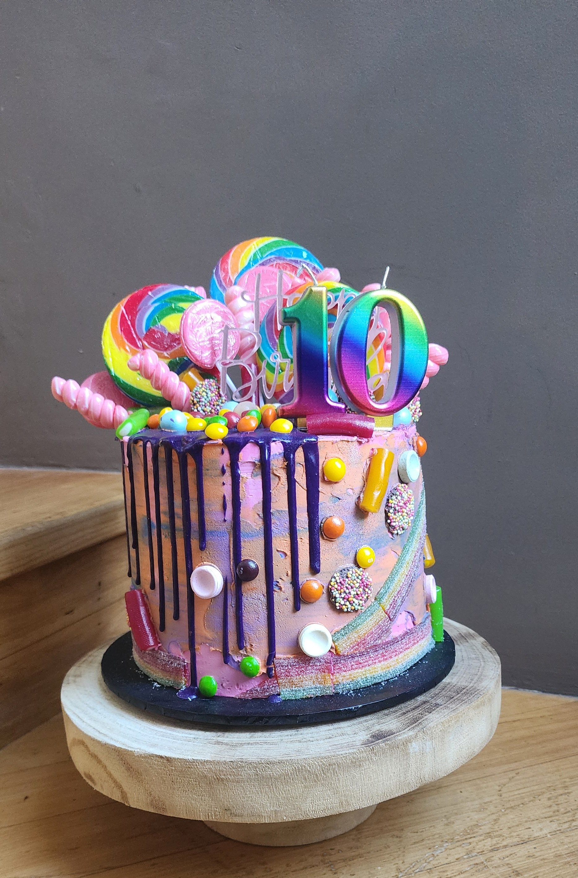 Colorful birthday cake decorated with rainbow lollipops, pink candies, and rainbow sprinkles, with a rainbow number 10 candle on top. The cake has purple and pink icing with a drip design, placed on a wooden cake stand.