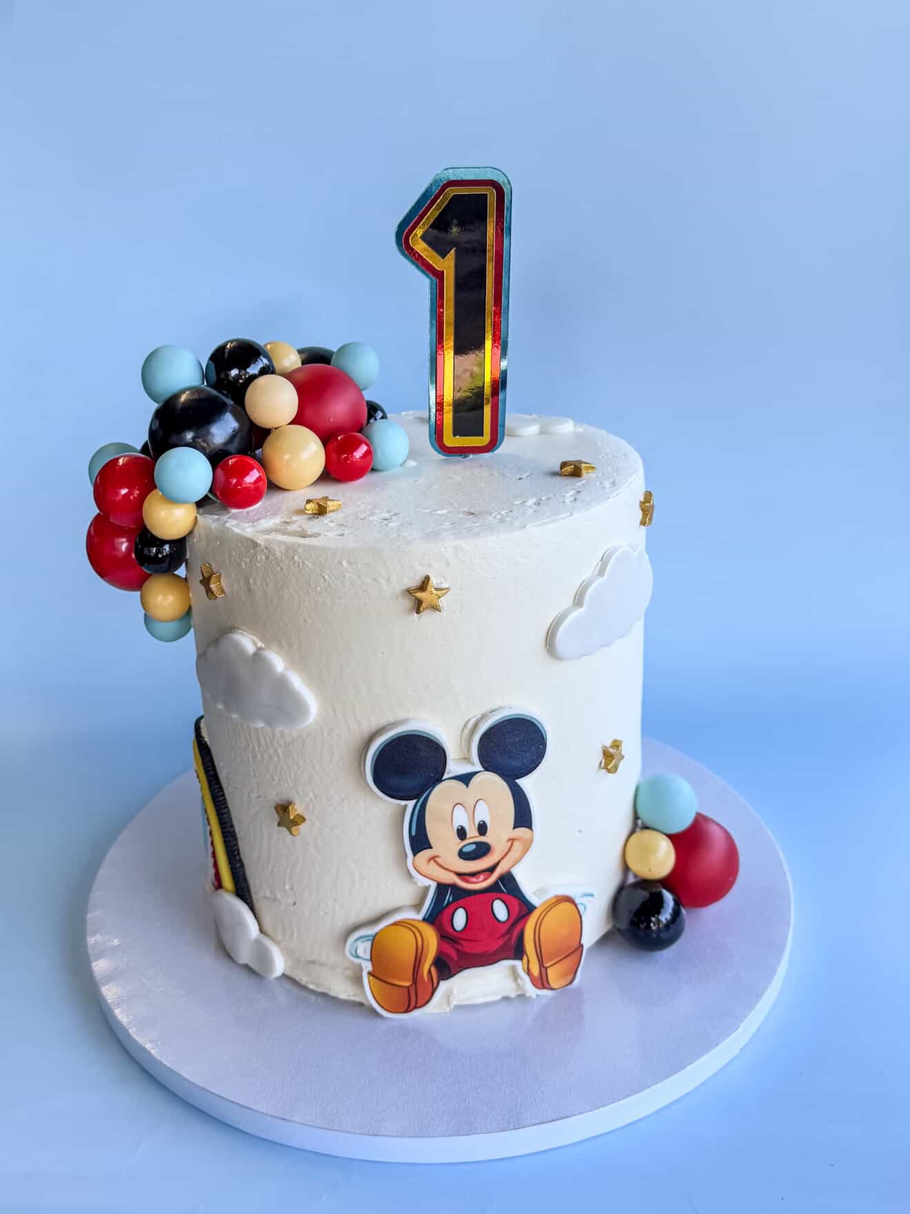 A birthday cake decorated with Mickey Mouse theme, featuring merging colorful balls, a number one candle, and Mickey Mouse images against a blue background.