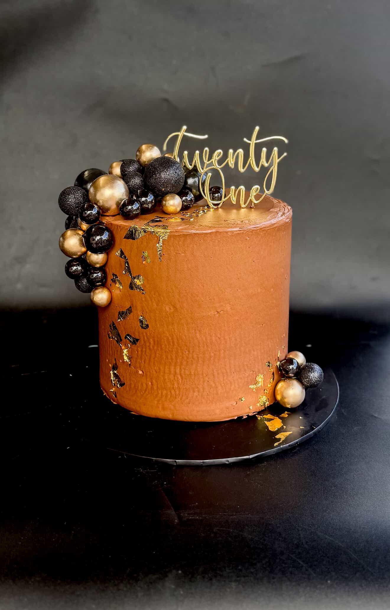 Chocolate birthday cake decorated with black, gold, and glittery spherical ornaments and a gold "Twenty One" topper, set on a black surface.
