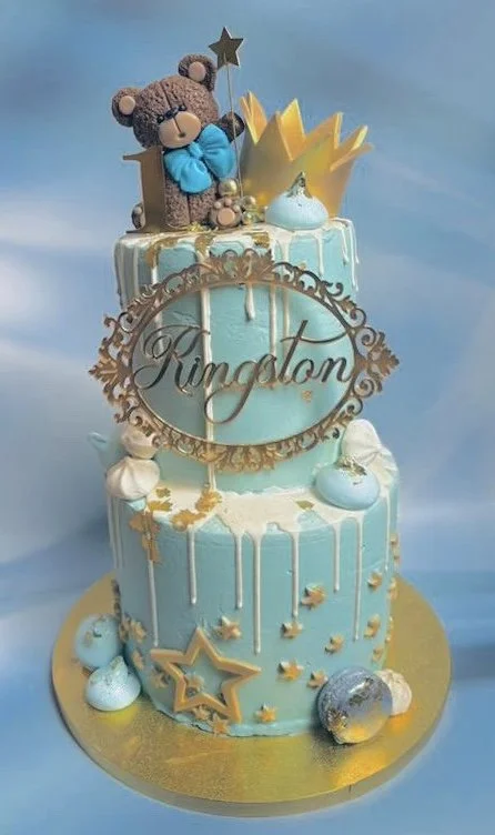 A two-tier baby shower cake in blue and white with a teddy bear topper, a gold crown, stars, and a personalized "Kingston" cake topper. Decorated with gold and white accents, the cake is on a gold cake board.