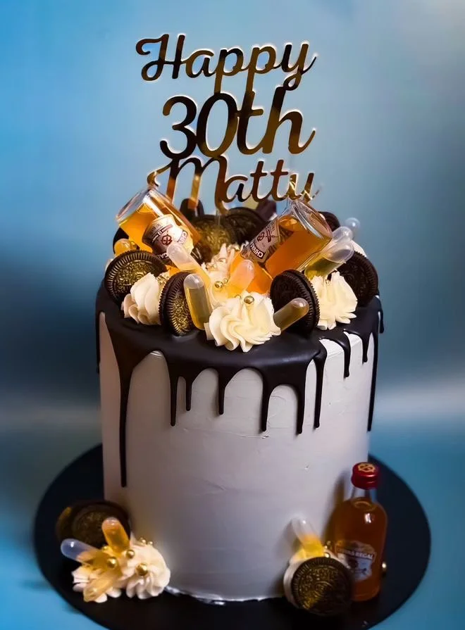 A birthday cake with a black and white theme, decorated with Oreo cookies, mini bottles of alcohol, and whipped cream. The cake features a black drip frosting and a gold-colored topper that reads 'Happy 30th Matty'.
