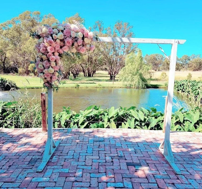 A floral arch decorated with pink and white roses and greenery, set outdoors by a pond with trees and greenery in the background under a clear blue sky.