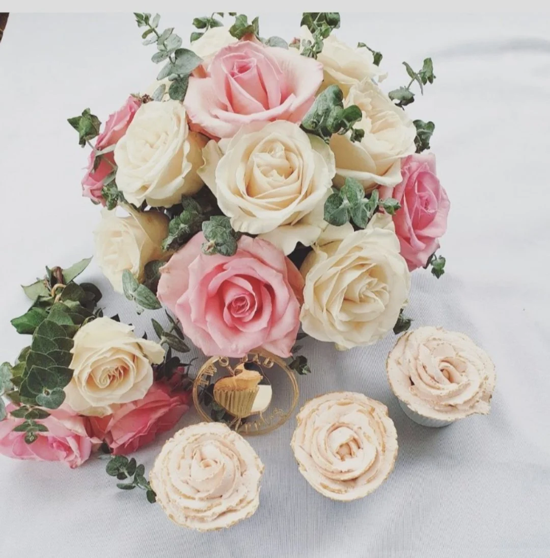 A bouquet of pink and cream roses with green foliage, accompanied by three cream-colored desserts topped with rose-shaped decorations, on a white surface.