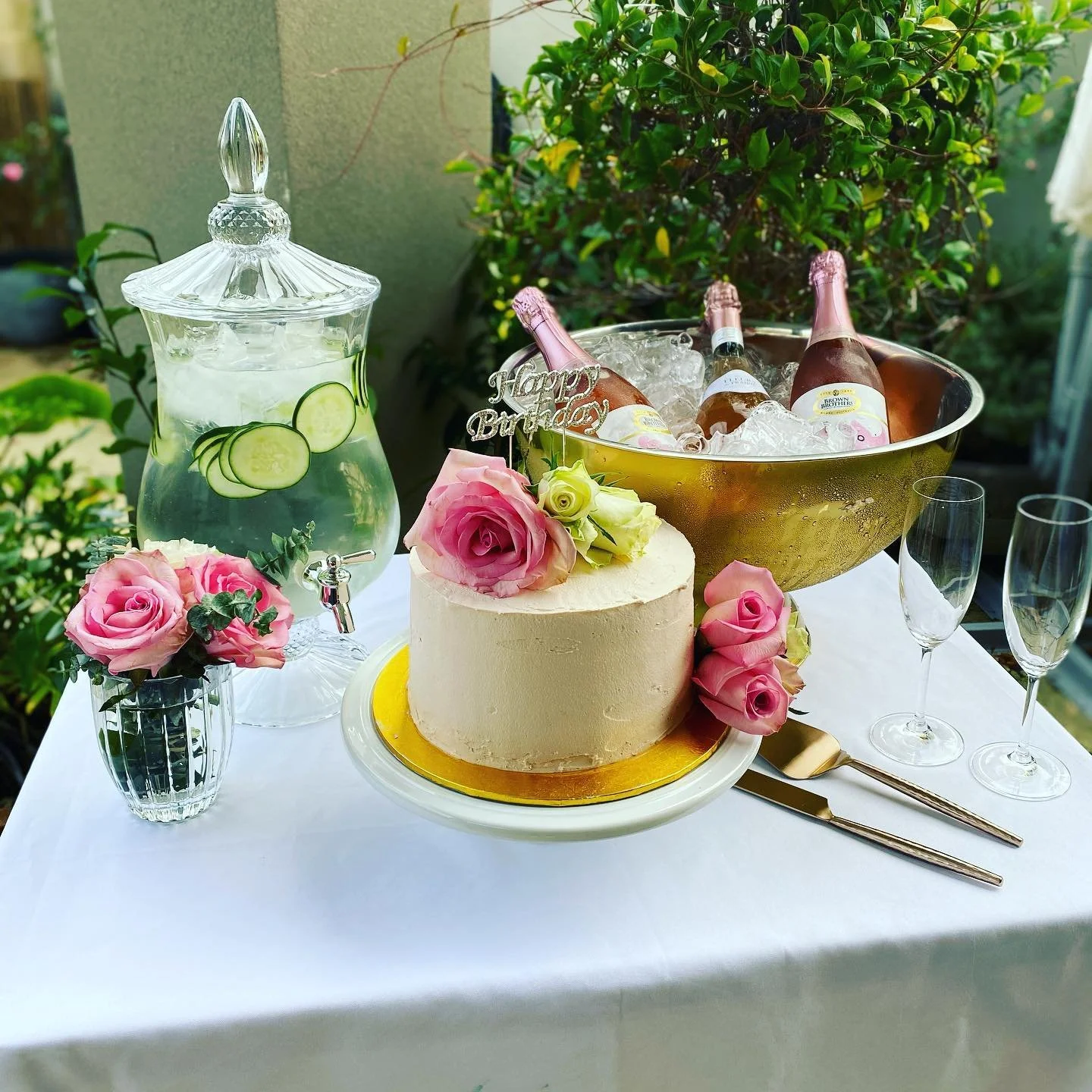 A birthday celebration setup with a white frosted cake decorated with pink and white roses, a glass beverage dispenser with cucumber water, a bowl of ice with bottles of pink champagne, empty champagne flutes, and a gold serving knife and cake server