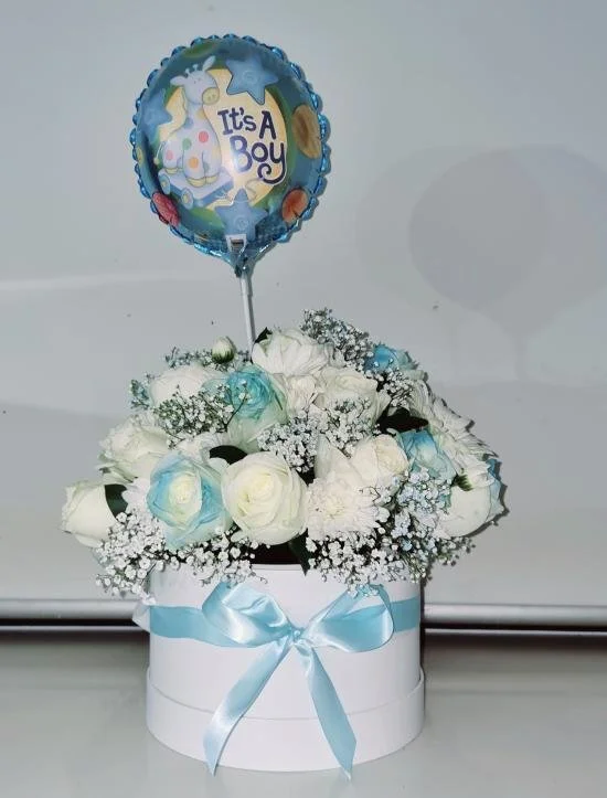 A white gift box with a blue ribbon bow contains white and light blue roses and baby's breath flowers. A blue helium balloon with baby shower themes floats above the flowers, saying "It's a Boy."