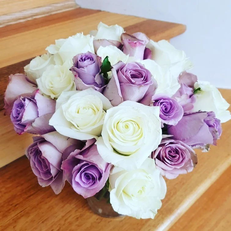 Bouquet of white, purple, and lavender roses in a glass vase on a wooden surface.