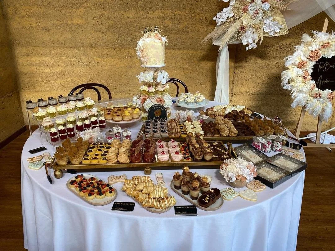 A dessert table with various pastries, cupcakes, cookies, and a tiered cake decorated with flowers, set for a celebration or wedding reception.