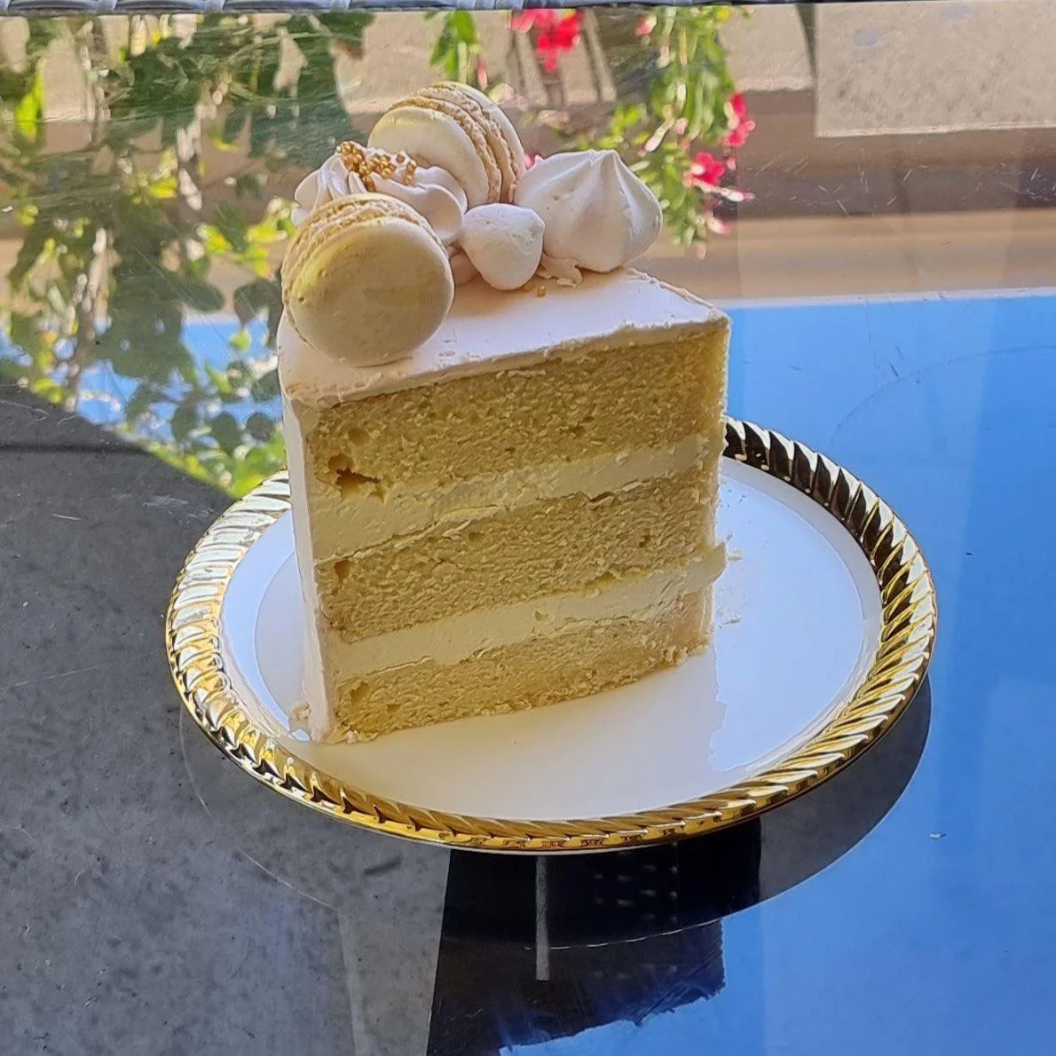 A slice of layered vanilla cake with white frosting, topped with meringue, macarons, and miniature marshmallows on a decorative gold-edged plate.