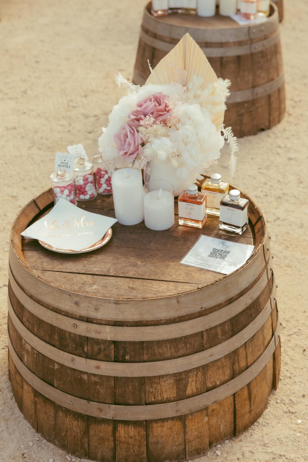 A wooden barrel used as a table with a floral arrangement, candles, perfume bottles, small jars of pink and white candies, and a plate with a napkin, in a rustic setting.