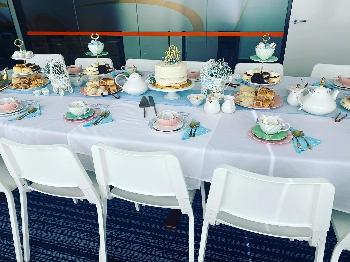 A decorated table set for a celebration with a white tablecloth, pastel-colored tea cups and plates, gold cutlery, a white cake with a "Oh Baby" topper, various pastries, a teapot, and floral arrangements.