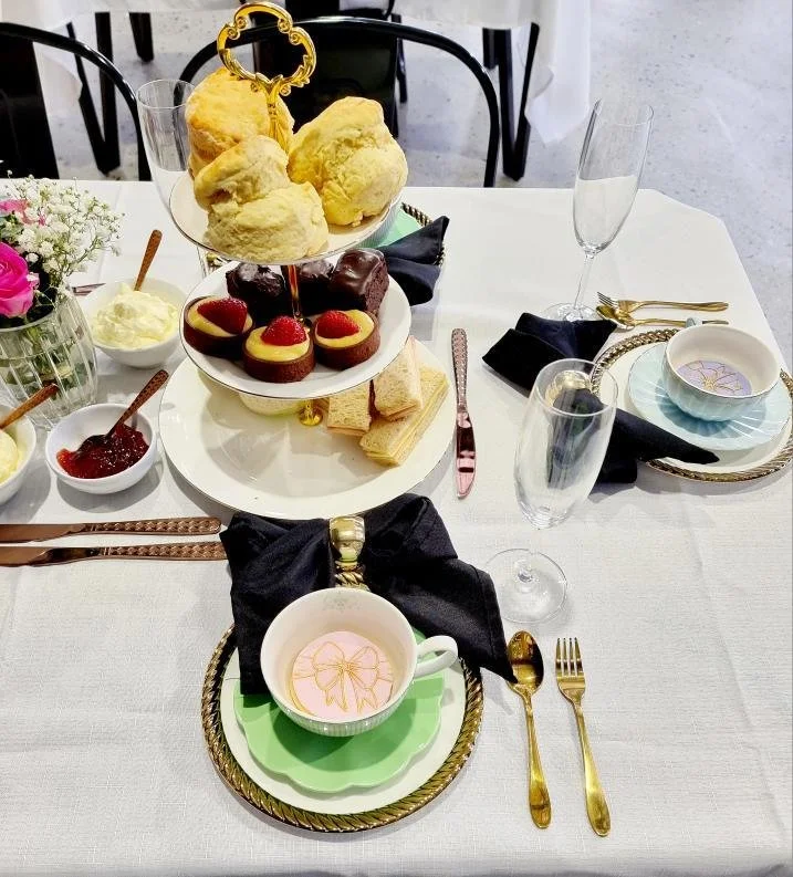 Table set for a formal afternoon tea with scones, strawberries, cake, and sandwiches, decorated with flowers and gold accents.
