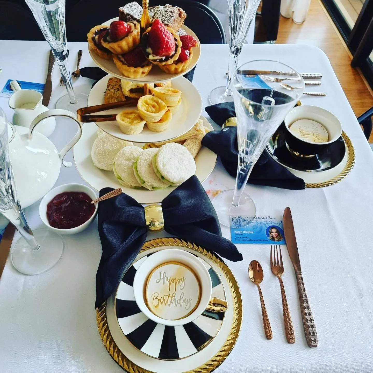 A birthday breakfast table with assorted pastries, sandwiches, a coffee cup with 'Happy Birthday' written inside, a small bowl of jam, and a card with a photo and name, set on a white tablecloth with black and gold decorations.