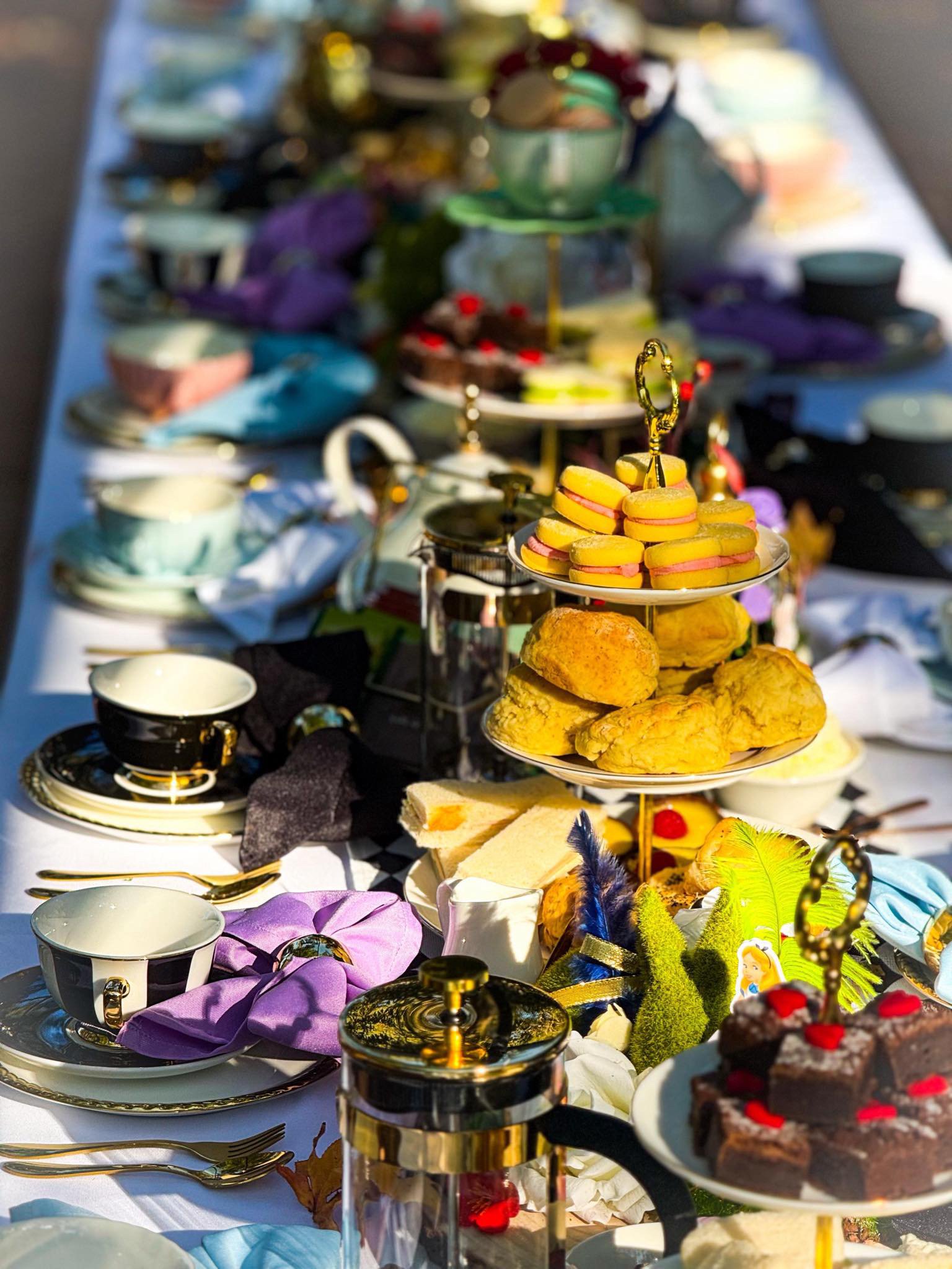 A table set for tea with various desserts, including macarons, scones, and cake, along with elegant cups, saucers, and utensils. The table is decorated with colorful feathers and flowers.