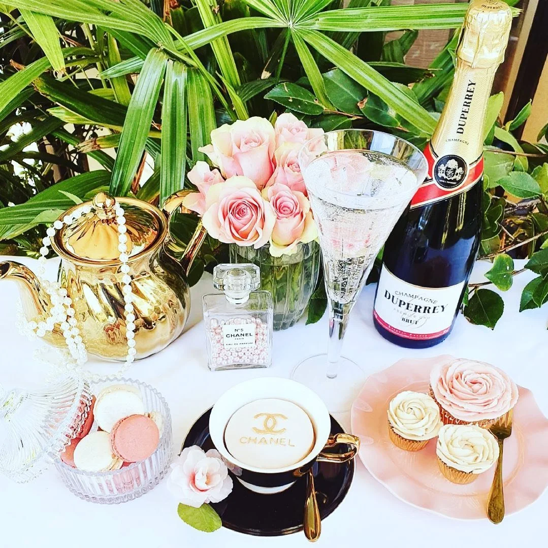 Elegant table setting with pink roses, a champagne bottle, a glass of champagne, a gold teapot, a Chanel perfume bottle, macarons, cupcakes with rose decorations, and champagne themed accessories, surrounded by lush green plants.