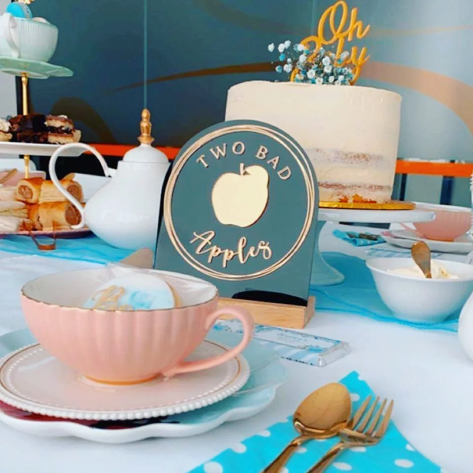 Table set with pink teacup and saucer, bowls of desserts, a sign reading 'Two Bad Apples' with an apple graphic, a white teapot, a marble cake with a 'Oh Baby' topper, and various plates and utensils.