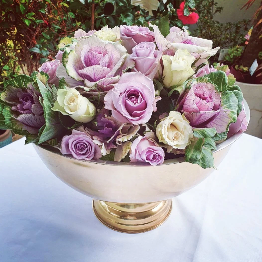 A decorative bowl filled with a variety of pink, purple, and white roses and ornamental cabbages, placed on a white surface with greenery in the background.