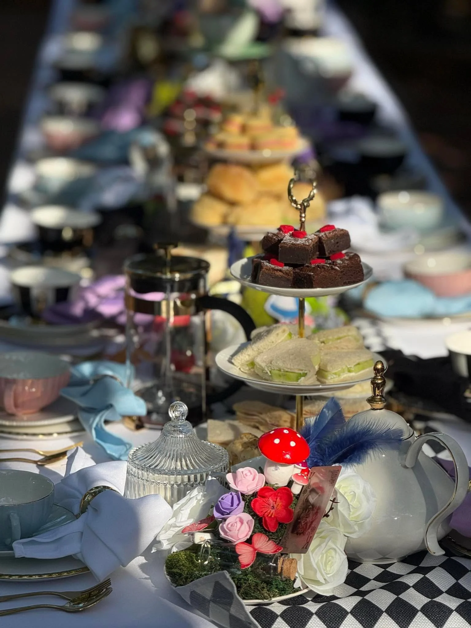 A dessert table with various sweet treats, including brownies with red sprinkles on a tiered tray, and finger sandwiches on a separate plate. The table is decorated with colorful artificial flowers, feathers, and a red mushroom ornament, with tea cup