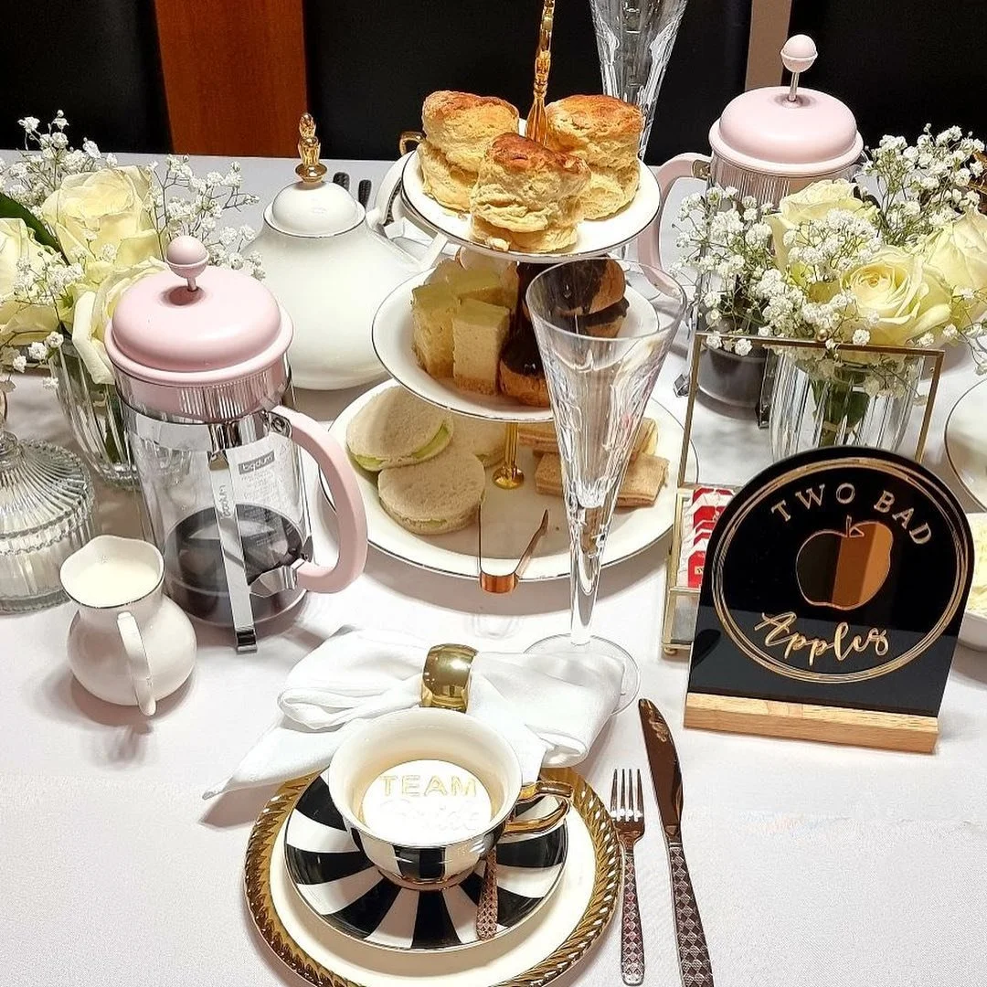Elegant table setting for afternoon tea with a three-tier tray of scones, sandwiches, and pastries, surrounded by flowers, teapots, and cups in black, white, and pink decor.