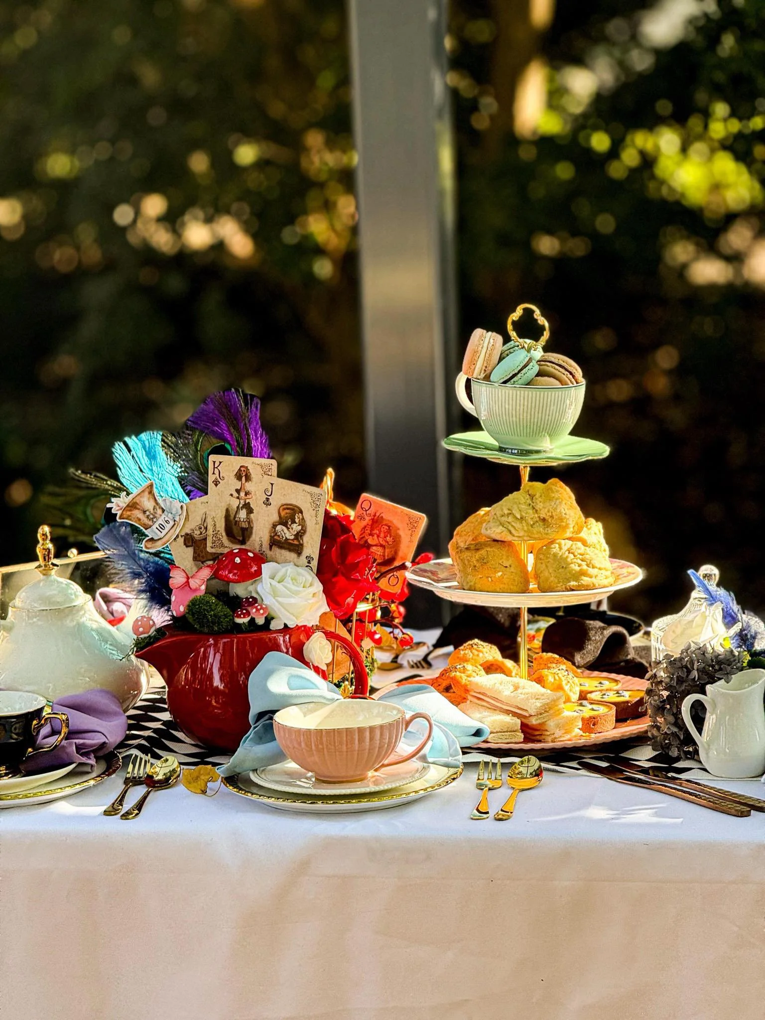 A table set for tea with plates, cups, and various desserts including macarons, cookies, and pastries. There are decorative elements such as playing cards, flowers, and feathers, with a tiered stand holding assorted treats. The background is blurred 