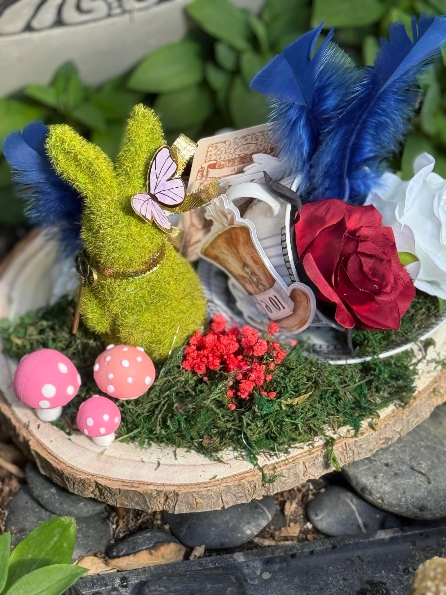 Decorative arrangement on a wooden slab with pink mushrooms, a yellow-green fuzzy bunny figure with a pink butterfly on its ear, red and white flowers, a blue feather, a white flower, and a paper with printed currency images, surrounded by greenery a