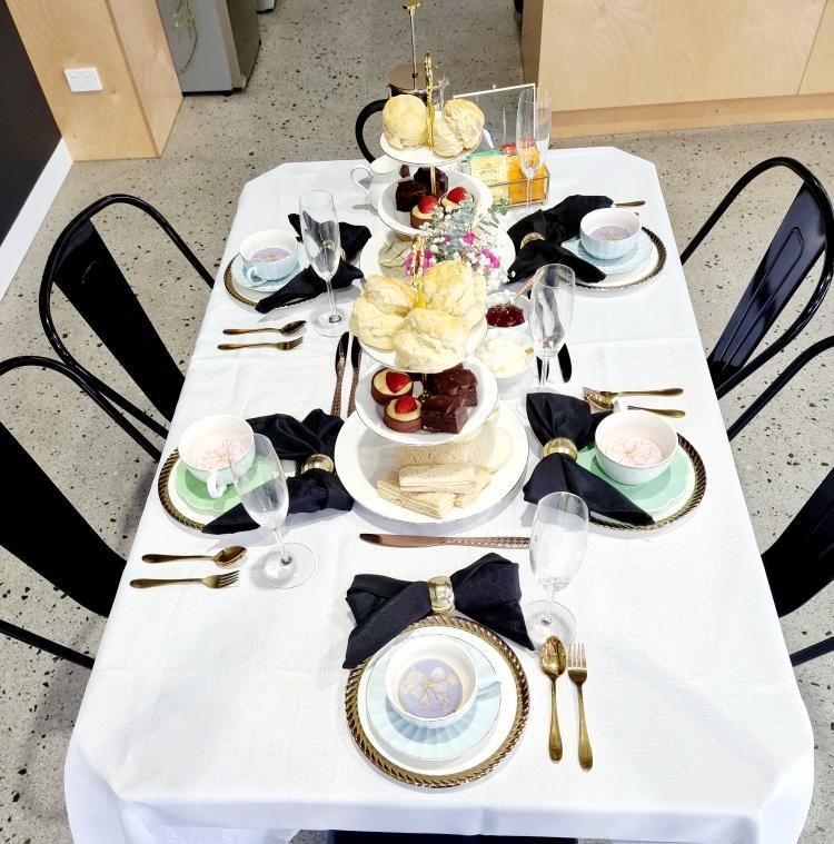 A formal dining table set for six with white tablecloth, black napkins held with gold napkin rings, gold utensils, clear champagne flutes, and pastel-colored plates. The table has a three-tiered serving tray with assorted desserts, including cream pu