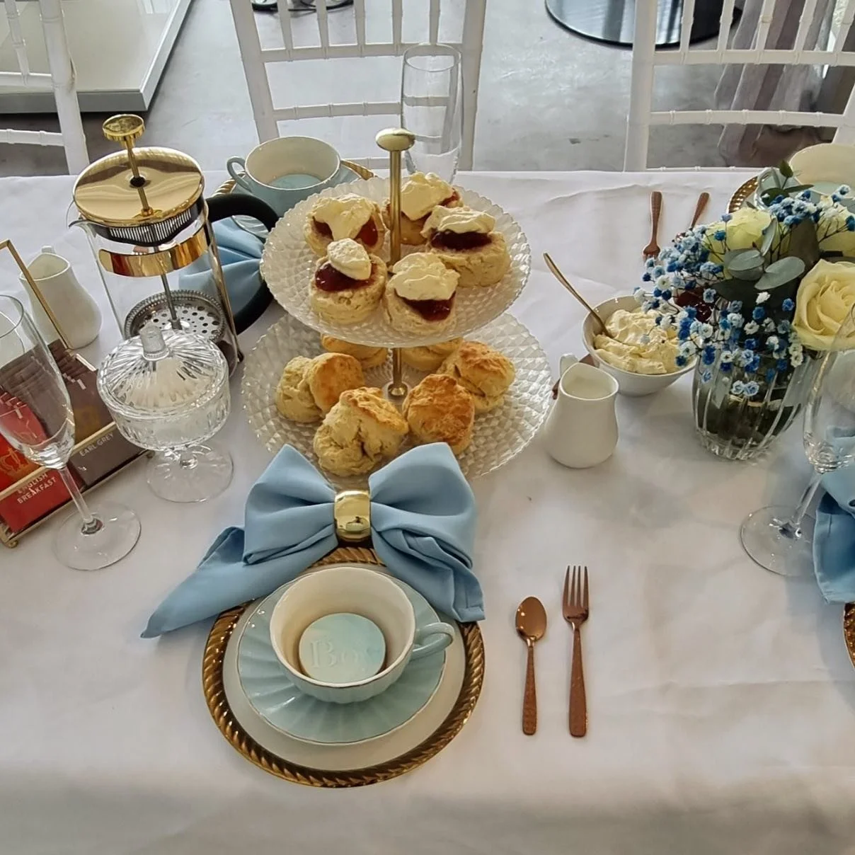 A table set with tea, scones, and pastries, decorated with a blue bow, flowers, and elegant tableware.