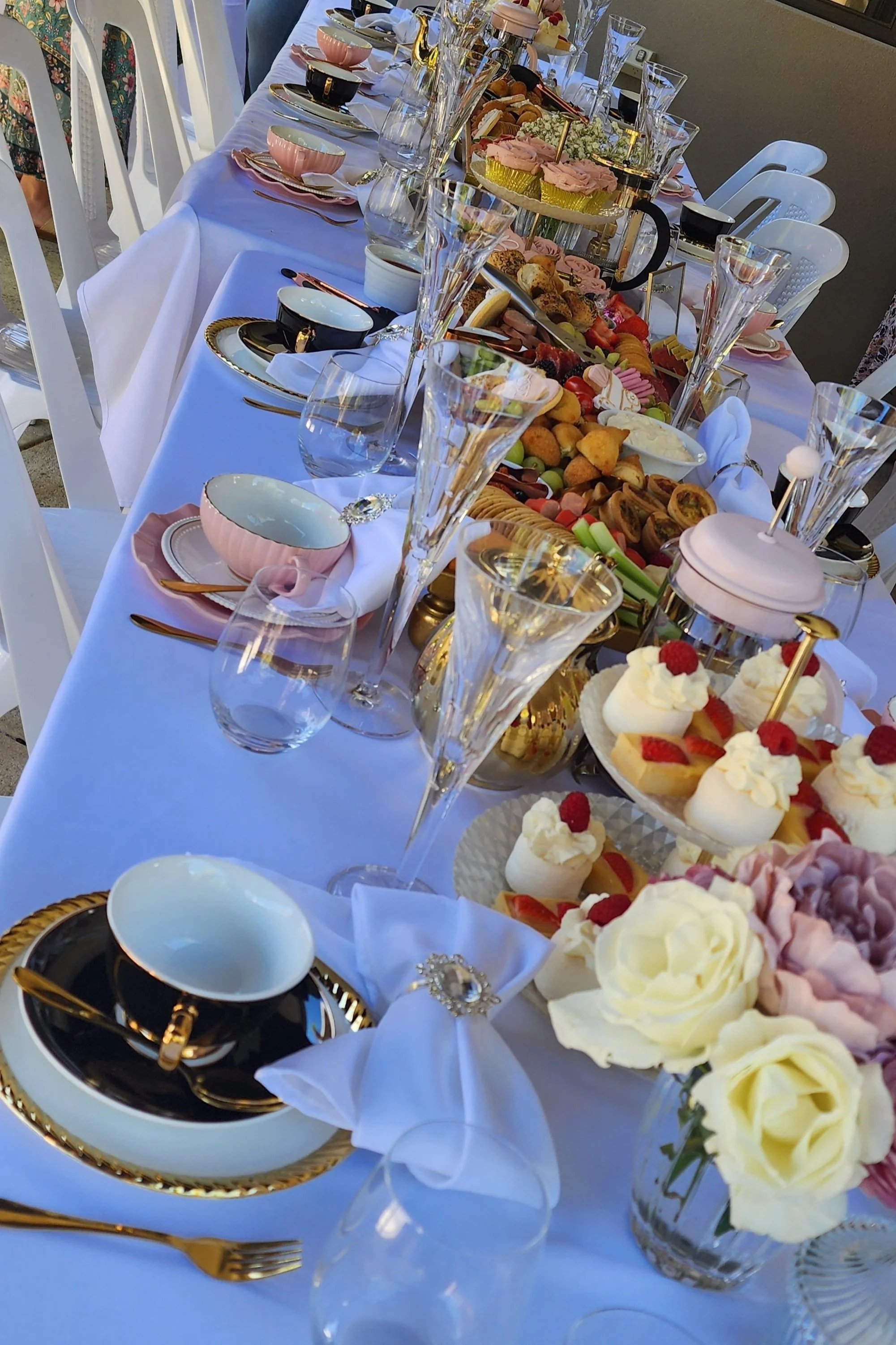 Elegant table set with white tablecloth, gold-rimmed china, gold flatware, and crystal glassware. Centerpieces include colorful flowers and a variety of desserts, fruits, and pastries. Pink, black, and white cups are placed on the table, and a tiered
