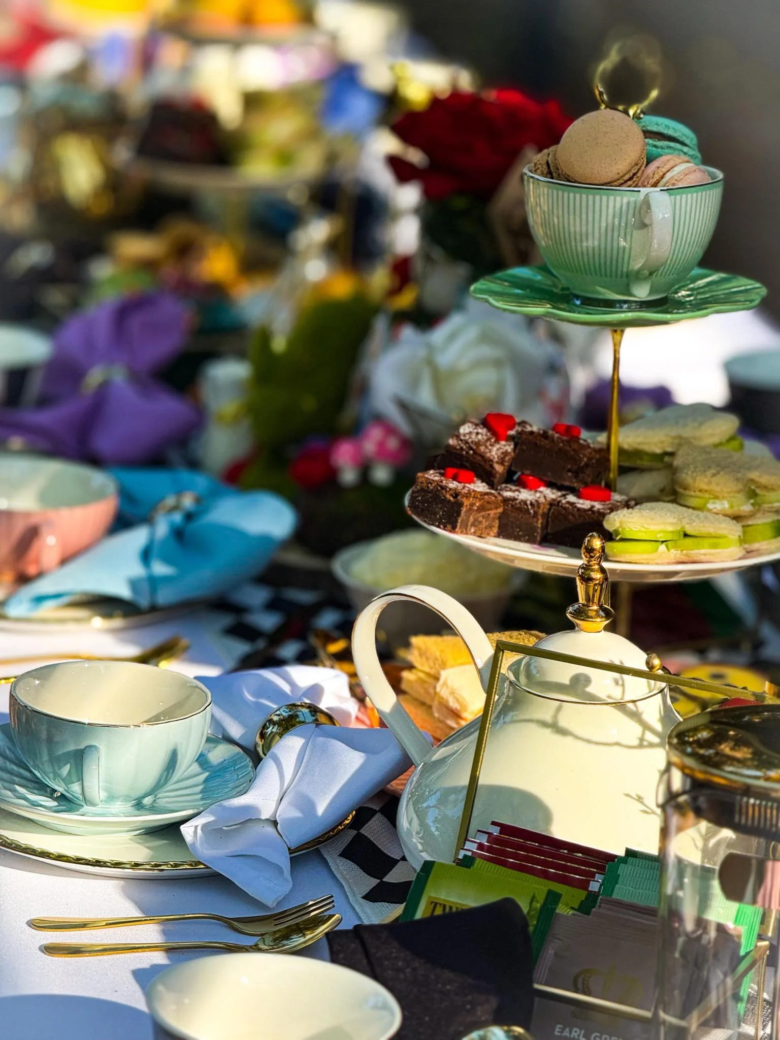 A table set with tea cups, saucers, and gold-colored cutlery, with a tiered tray of macarons and other desserts, and various packages and items in the background.