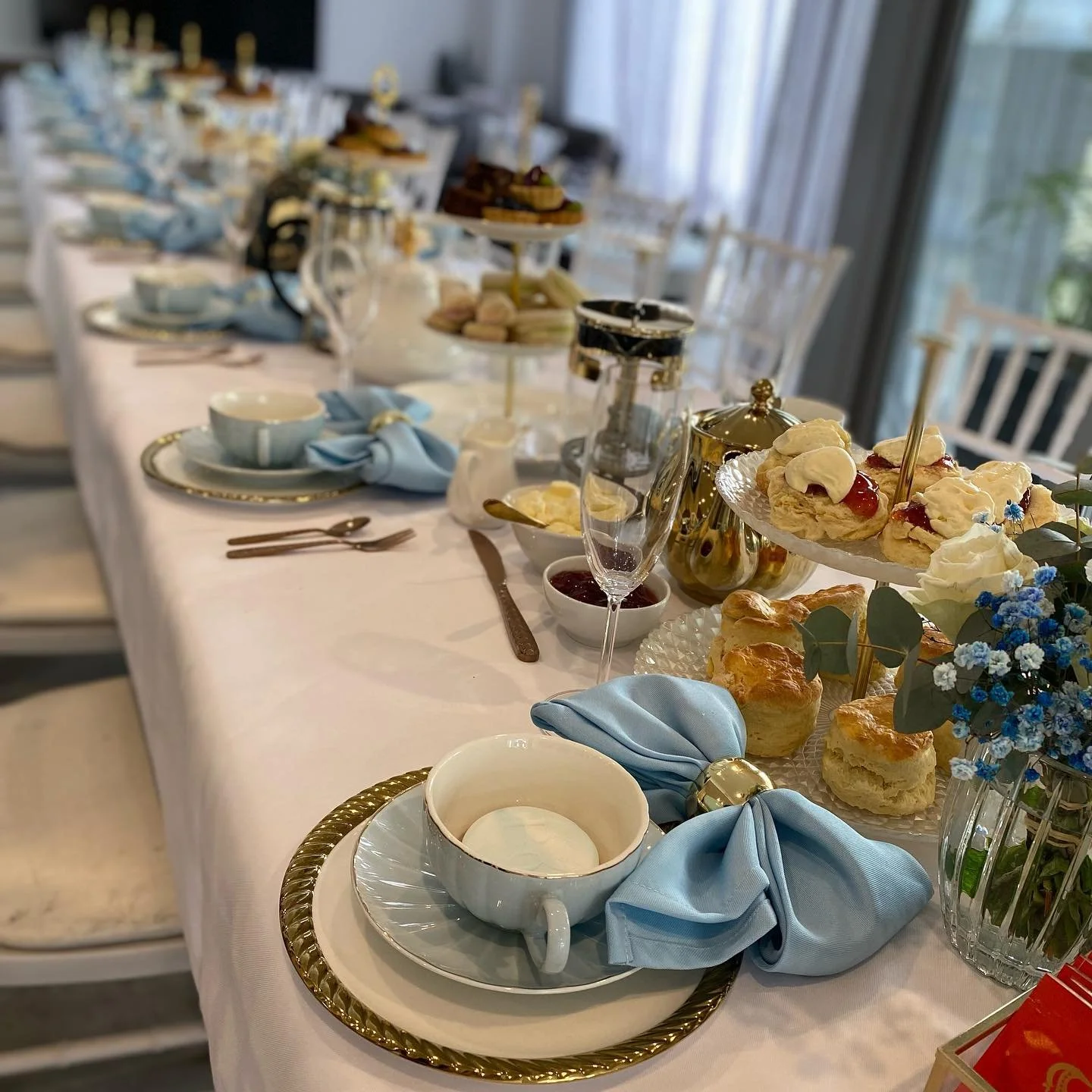 Elegant table setup for afternoon tea with china cups, gold-rimmed plates, blue napkins, assorted pastries, scones, sandwiches, and floral decorations, with a long white table and chairs in the background.