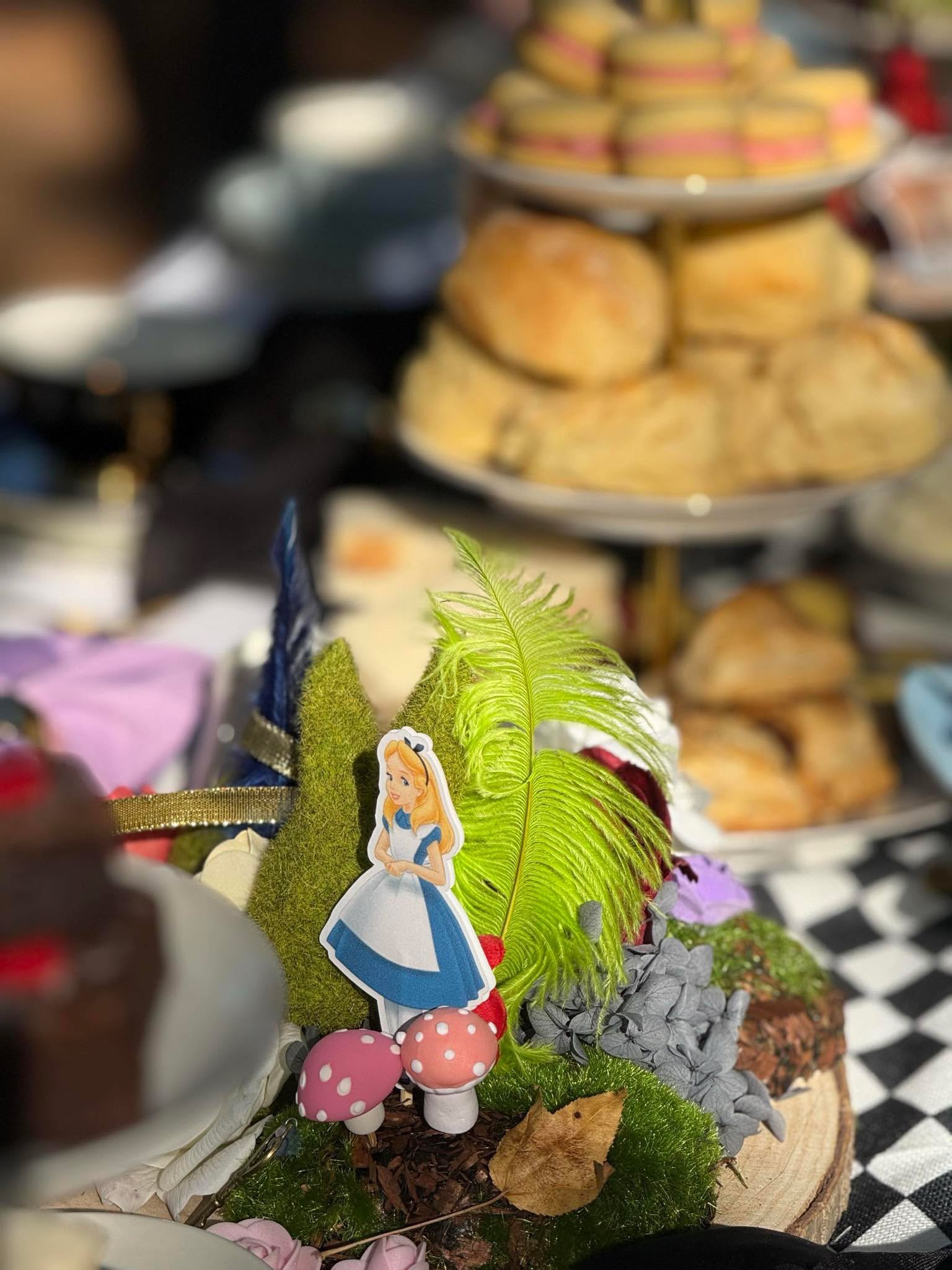 Decorative scene with a Disney Princess cutout of Alice from Alice in Wonderland, pink mushrooms with white spots, green moss, dried leaves, and artificial plants arranged on a wooden surface.