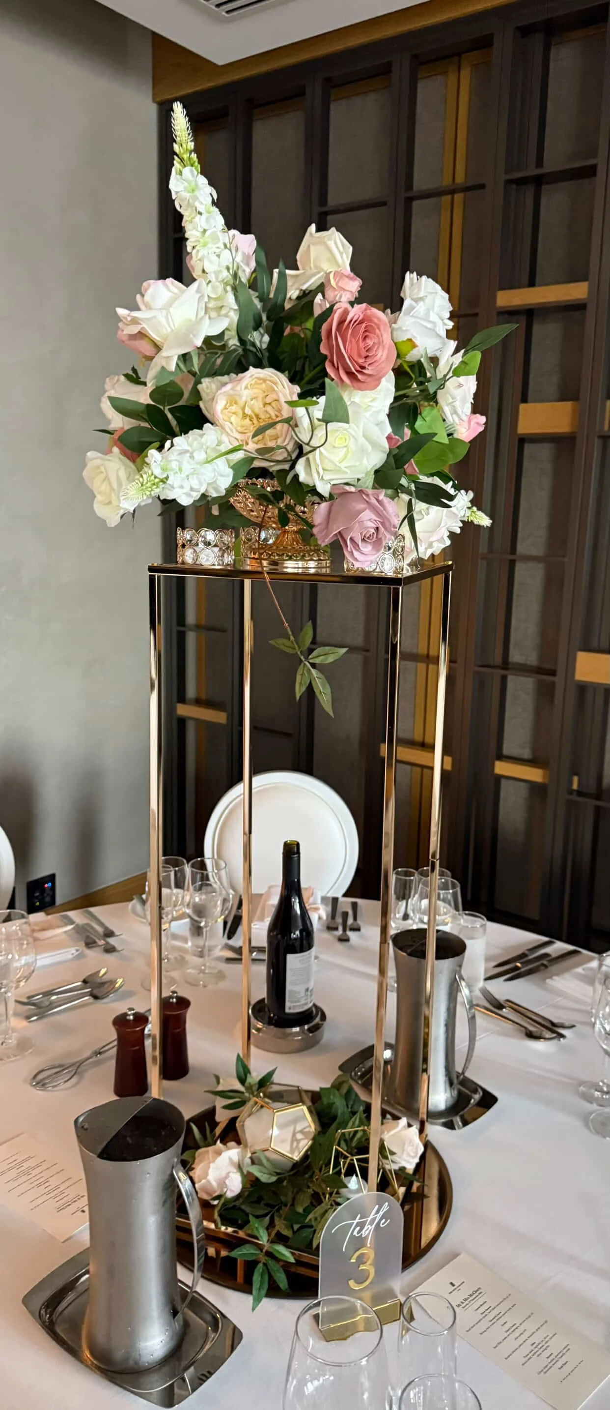 Elegant table centerpiece with a tall floral arrangement of pink and white roses and other flowers, placed on a gold and glass stand. The table is set with wine glasses, silverware, a bottle of wine, salt and pepper shakers, and a decorative tray wit