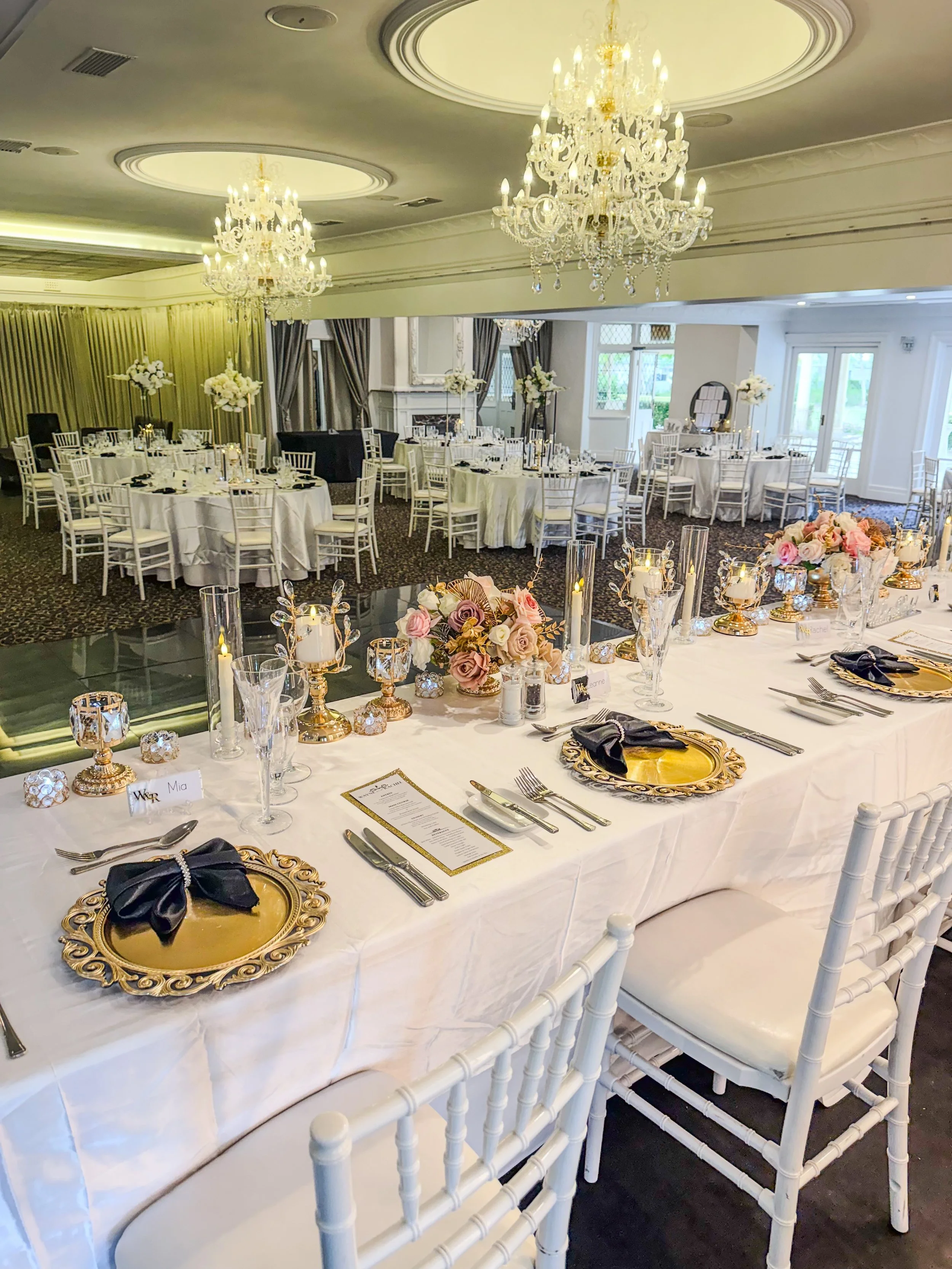Elegant banquet hall decorated for a wedding reception with round tables draped in white tablecloths, floral centerpieces, candles, and ornate gold-rimmed plates and black napkins. Two large crystal chandeliers hang from the ceiling, and white chairs