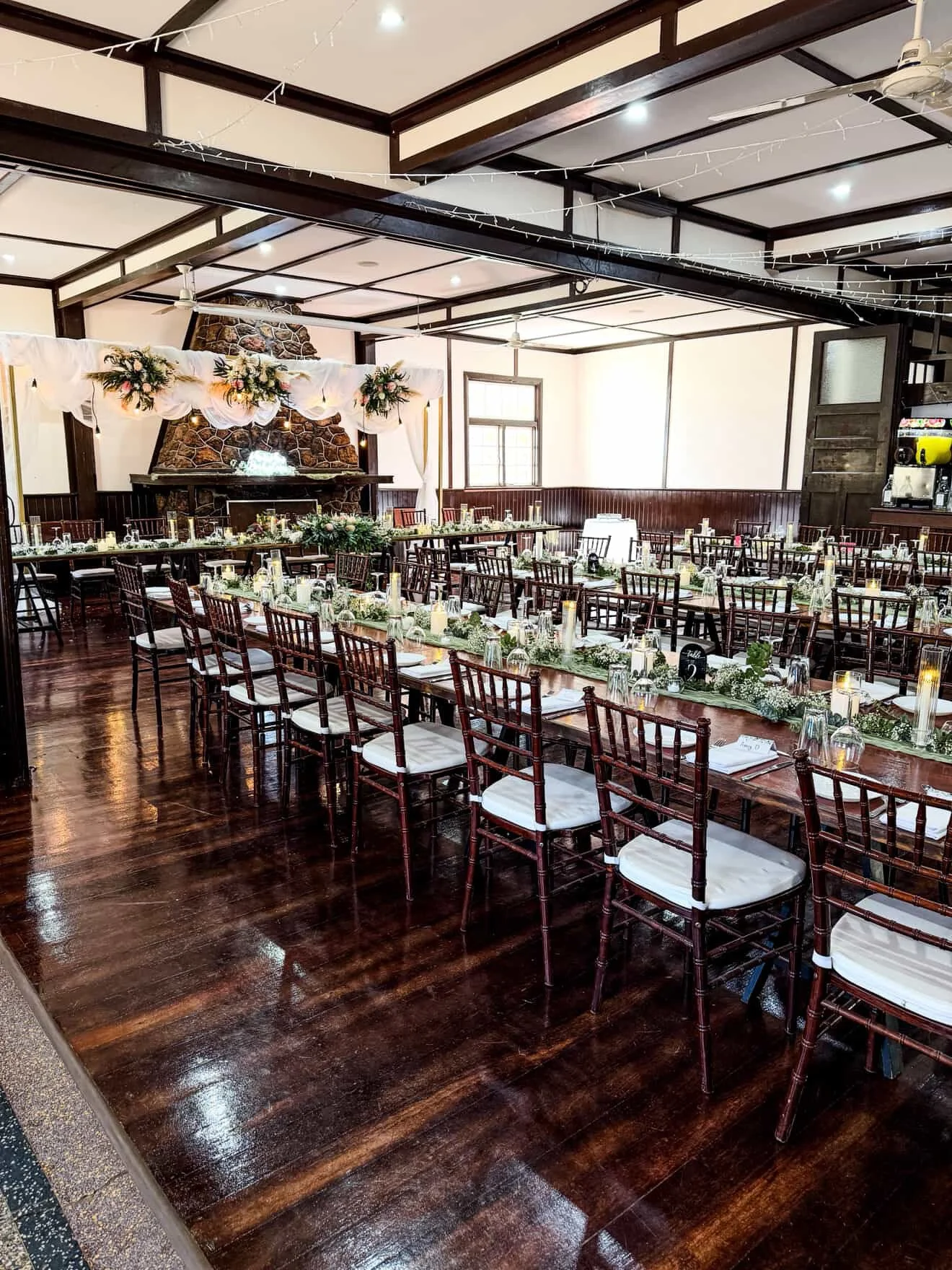 Indoor event space decorated for a wedding reception with long tables, floral centerpieces, candles, and a decorated fireplace on a wood-paneled wall.