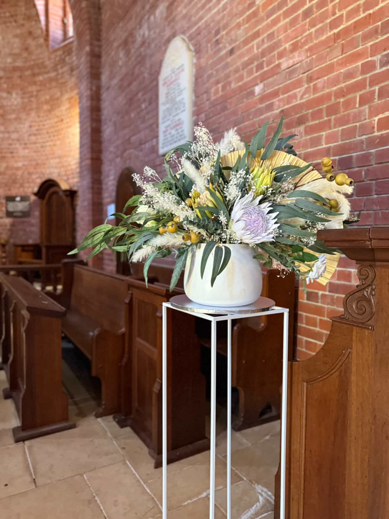 Large floral arrangement with white, yellow, and purple flowers and greenery in a white vase on a tall metal stand inside a brick-walled church.