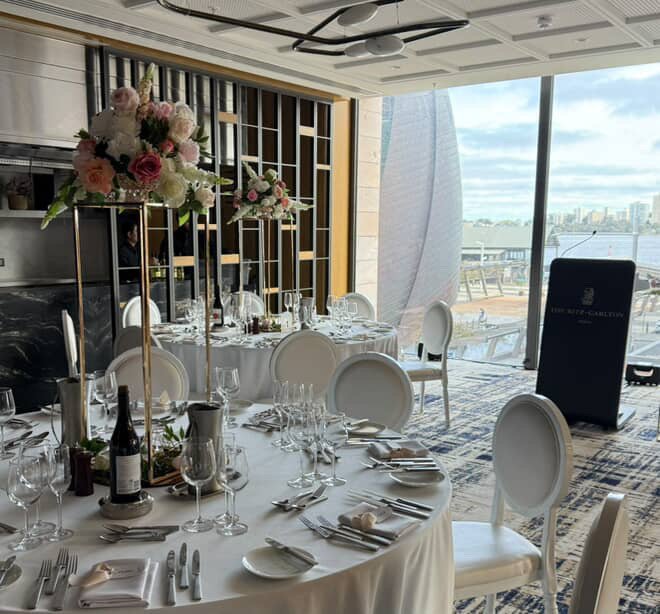 Elegant dining table set for a formal event with white tablecloths, floral centerpieces, wine bottles, and glassware, in a high-rise venue with city and waterfront view.