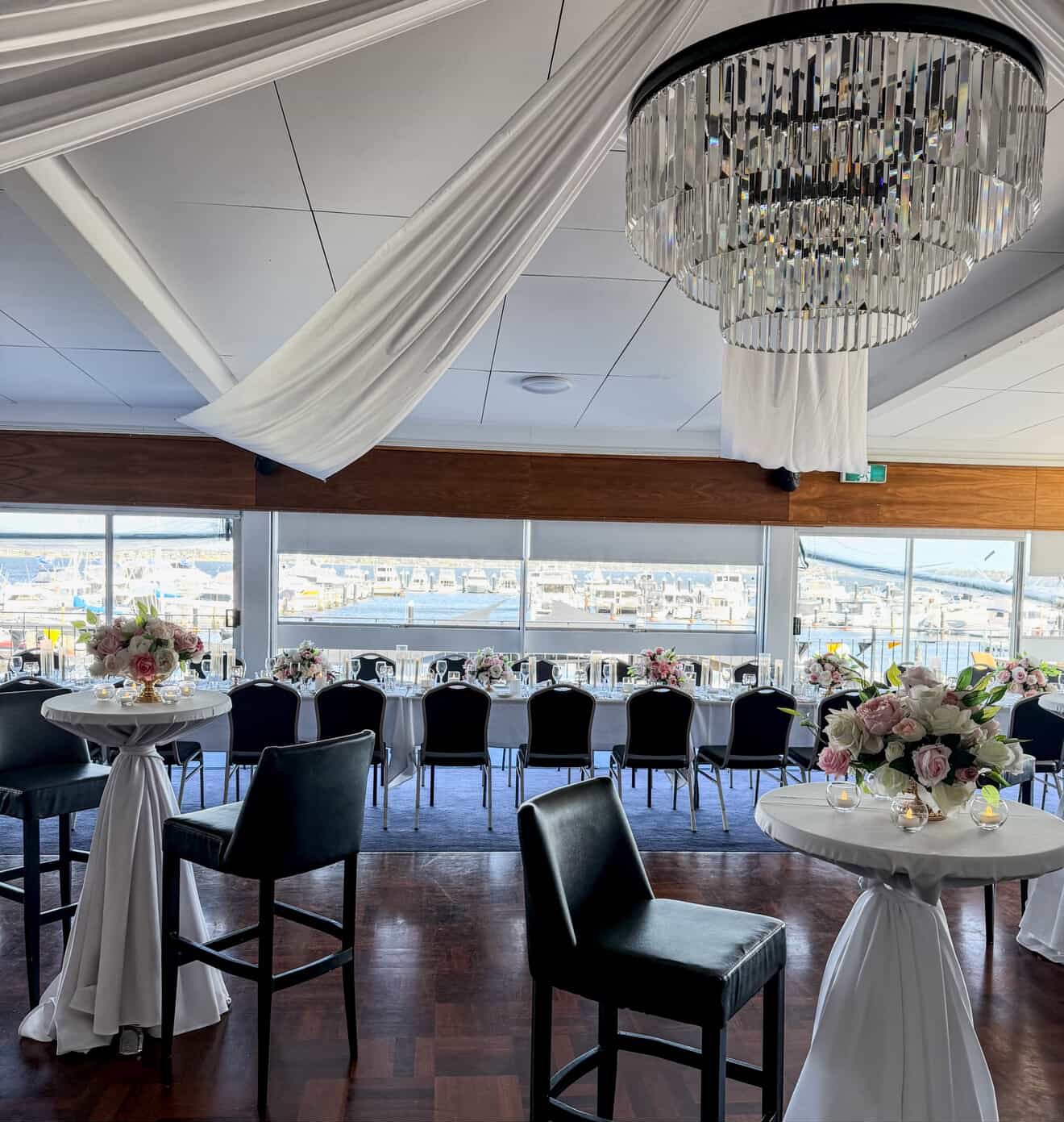 Elegant event space with round tables decorated with pink and white floral centerpieces, black chairs, a large window view of city buildings, and a chandelier hanging from the ceiling.
