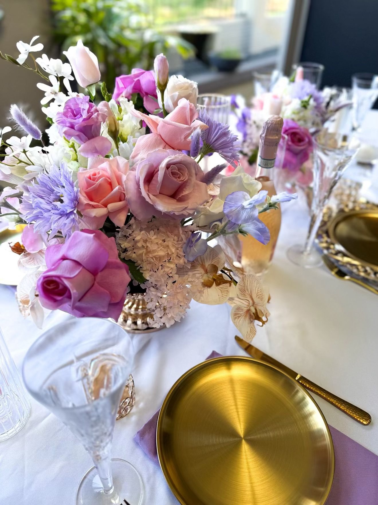 Elegant table setting featuring a floral centerpiece with pink, purple, and white flowers, gold-rimmed plates, gold cutlery, clear glassware, and a white tablecloth, illuminated by natural light.